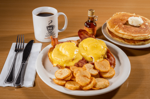 A plate of eggs Benedict with bacon, crispy potatoes, pancakes, and a cup of coffee on a wooden table.