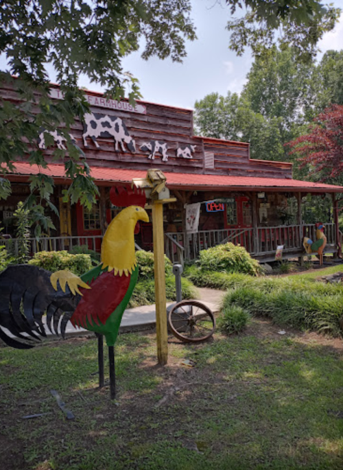 A rustic farmhouse with a red roof, surrounded by greenery and a colorful rooster statue in front.