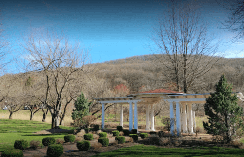 A serene park scene featuring a white pergola, bare trees, and a distant hill under a clear blue sky.