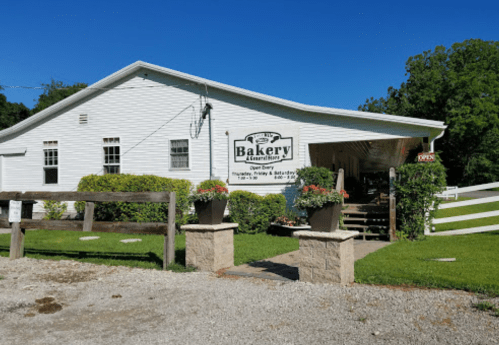 A charming white bakery building with a sign, surrounded by greenery and flower pots, on a sunny day.