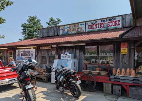 A rustic country store with wooden signage, parked motorcycles, and handmade crafts displayed outside.