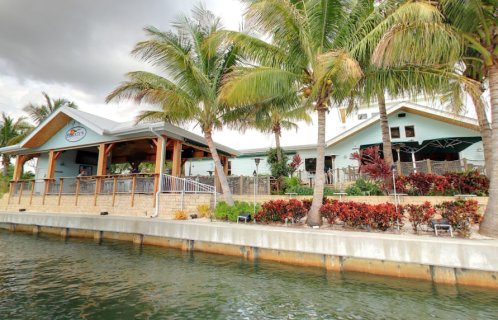 A waterfront restaurant surrounded by palm trees and colorful landscaping, with a cloudy sky above.