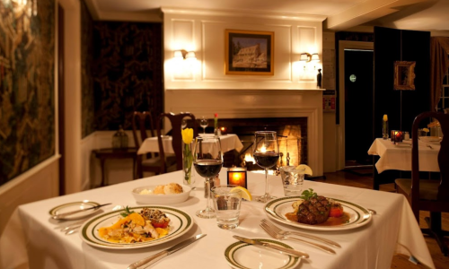 A cozy restaurant table set with gourmet dishes, wine glasses, and a warm fireplace in the background.