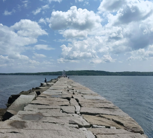 A rocky pier extends into the water under a cloudy sky, with a lighthouse visible in the distance.