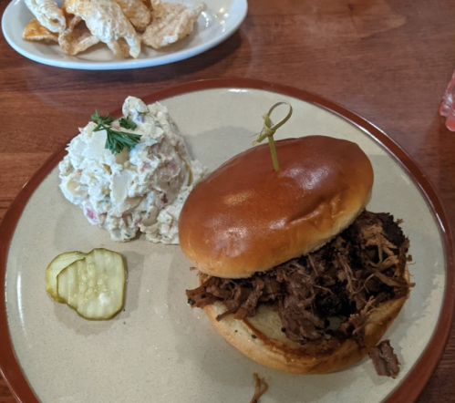 A pulled pork sandwich on a bun with a pickle slice, served alongside creamy potato salad.