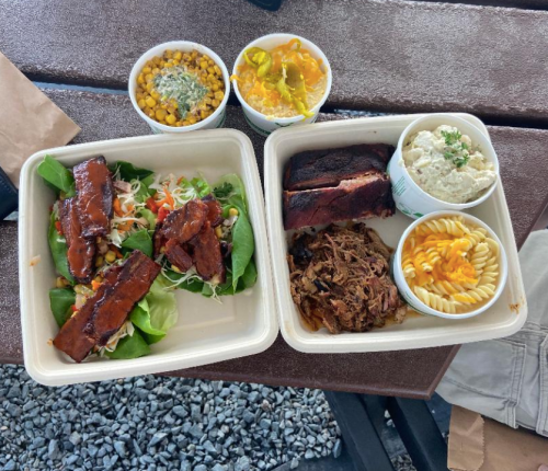 A takeout meal featuring BBQ ribs, pulled pork, macaroni, corn salad, and a colorful salad on a bed of greens.