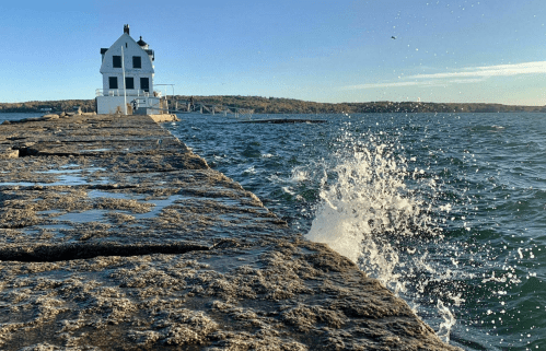 A rocky pier extends into the water, with a white lighthouse in the background and waves splashing against the rocks.