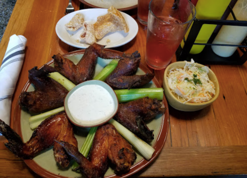 A platter of chicken wings arranged with celery sticks and a bowl of ranch dressing, alongside coleslaw and a drink.