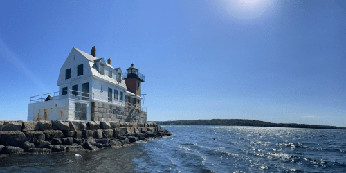 A white lighthouse and house on rocky shore, with calm blue waters and a clear sky in the background.