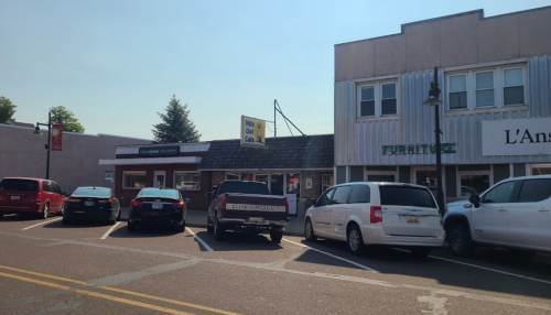 A street view of a small town with parked cars and storefronts, including a furniture shop and a café.