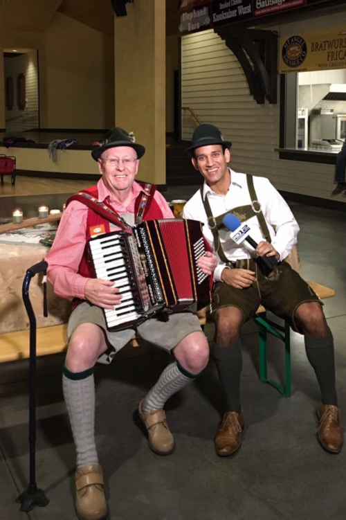 Two men in traditional German attire sit together, one playing an accordion, smiling at the camera.