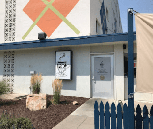 Exterior of a building with a sign reading "PAC" and a decorative mural, featuring plants and a blue fence.