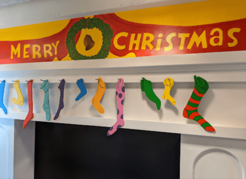 Colorful stockings hang on a mantel beneath a festive "Merry Christmas" sign with a wreath.