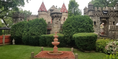 A stone castle with red-topped towers surrounded by lush greenery and a decorative fountain in the foreground.