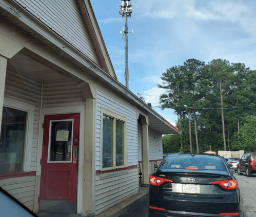 A drive-thru restaurant with a red door and a cell tower in the background, surrounded by trees and parked cars.
