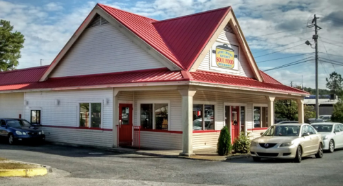 A cozy soul food restaurant with a red roof and white exterior, surrounded by parked cars and a blue sky.