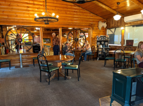 Interior of a rustic restaurant with wooden walls, tables, and large wheel decorations; patrons dining and chatting.