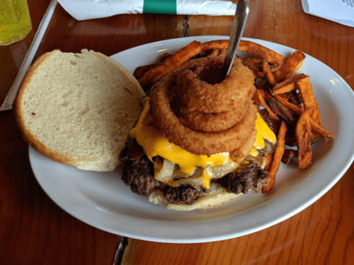 A hearty burger with melted cheese, onion rings on top, served with sweet potato fries on the side.