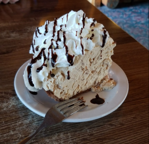 A slice of creamy pie topped with whipped cream and chocolate drizzle, served on a white plate with a fork.