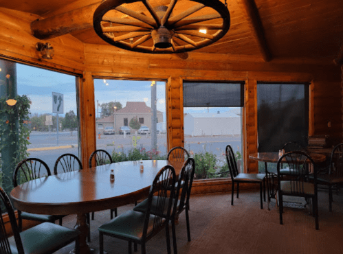 Cozy wooden dining area with a large round table and chairs, featuring large windows and a rustic ceiling fan.
