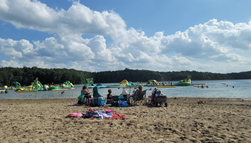 A sandy beach with people relaxing, colorful inflatable water toys in the background, and a clear blue sky.