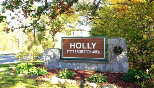 Sign for Holly State Recreation Area, surrounded by trees and greenery, with a clear blue sky above.