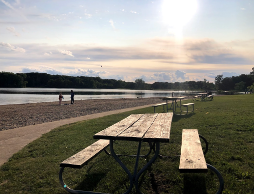 A serene lakeside scene with picnic tables, a sandy beach, and two people walking along the water under a sunny sky.