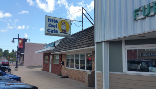 Sign for Nite Owl Cafe with a cartoon owl, located on a street with shops and clear blue skies.