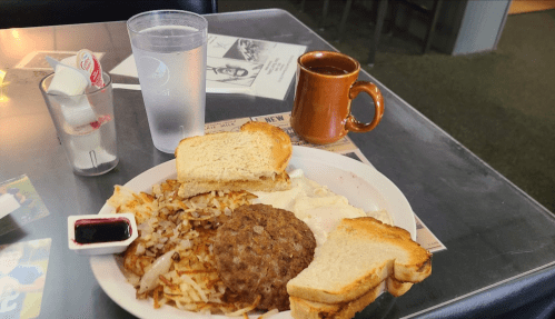 A plate of breakfast with eggs, hash browns, sausage patty, toast, and a side of syrup, with coffee and water.