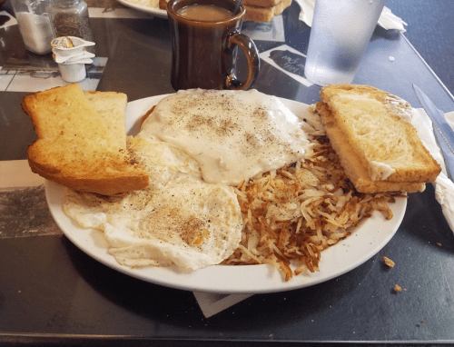 A plate of breakfast with two fried eggs, hash browns, toast, and a cup of coffee on a table.