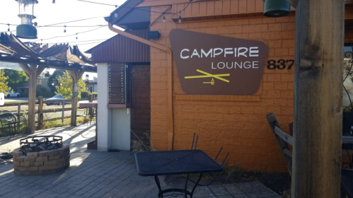 Exterior of Campfire Lounge with a sign, outdoor seating, and a fire pit in a sunny setting.