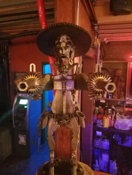 A whimsical metal sculpture of a mariachi figure with gears and a sombrero, set in a colorful bar environment.