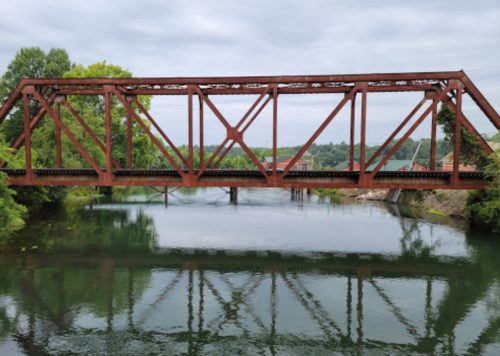 Rusty red railroad bridge over a calm river, surrounded by greenery and cloudy skies. Reflections visible in the water.