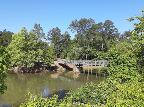 A metal bridge spans a calm pond, surrounded by lush green trees under a clear blue sky.