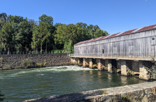 A wooden structure beside a river, surrounded by trees under a clear blue sky. Water flows gently beneath the building.