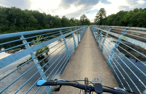 A bike view from a blue metal bridge over a calm river, surrounded by lush green trees under a cloudy sky.