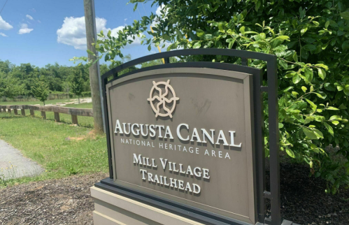 Sign for Augusta Canal National Heritage Area, Mill Village Trailhead, surrounded by greenery and blue sky.