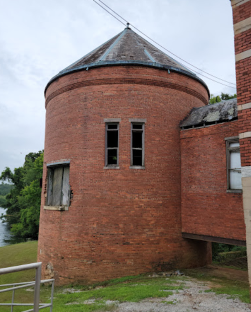 A round, brick building with a conical roof, featuring several windows and surrounded by greenery.