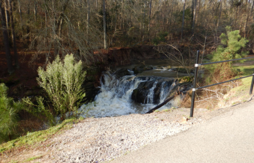 A small waterfall cascades over rocks, surrounded by trees and greenery, with a gravel path nearby.