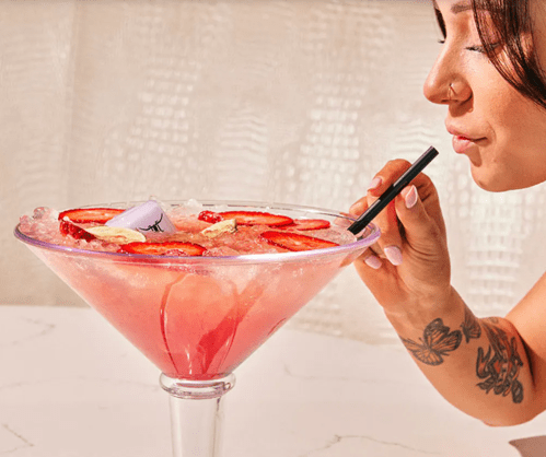 A person sipping a large pink cocktail garnished with strawberries and ice, using a black straw.