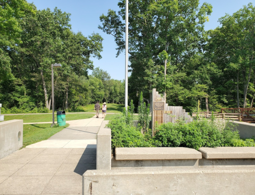 A sunny park path with trees on either side, leading to a wooden structure and two people walking in the distance.