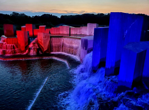 A colorful waterfall feature with illuminated geometric structures at sunset, reflecting vibrant hues in the water.