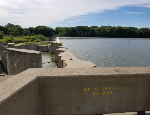 A concrete dam with a "No Climbing" sign, overlooking a calm lake and lush greenery under a blue sky.