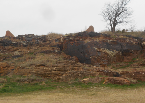 Rocky hillside with sparse grass, featuring a few prominent rock formations and a solitary tree in the background.