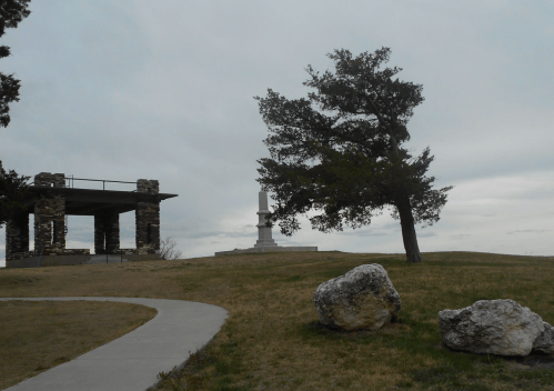 A stone structure and a tall monument stand on a grassy hill under a cloudy sky, with a tree nearby.