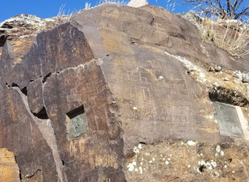 Ancient petroglyphs carved into a rocky surface, surrounded by sparse vegetation and a clear blue sky.