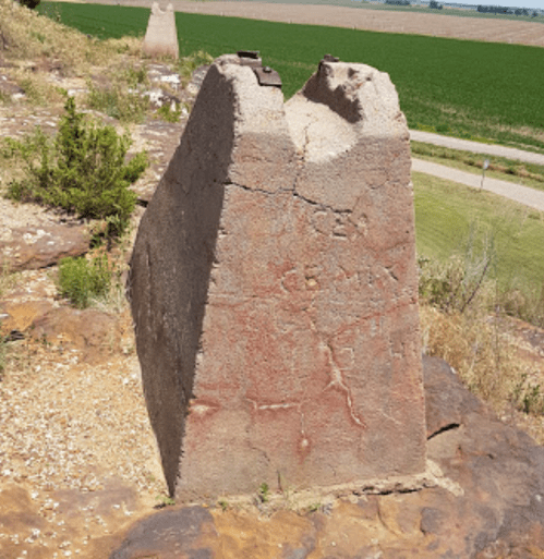 Weathered stone monument with inscriptions, set against a backdrop of green fields and a dirt road.