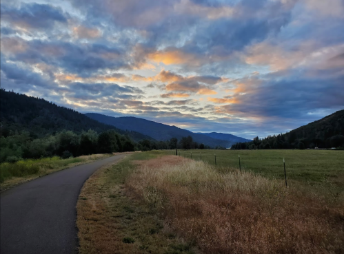 A winding path through a grassy field, with mountains and a colorful sunset sky in the background.