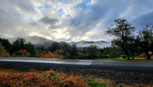 A scenic view of misty mountains under a cloudy sky, with colorful autumn trees lining the roadside.