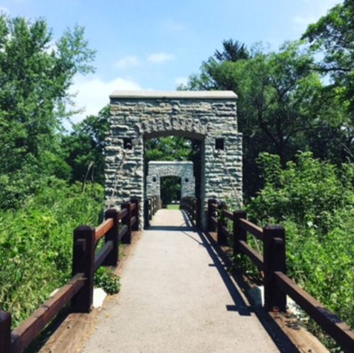 A stone archway leads into a lush green pathway, surrounded by trees and foliage under a clear blue sky.